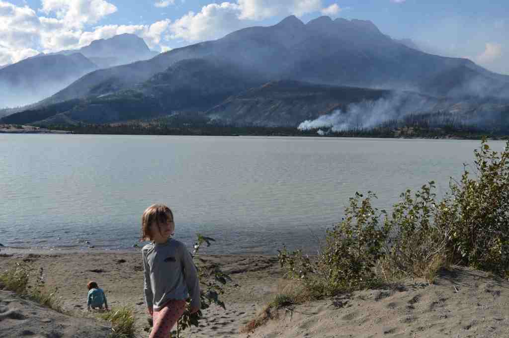 Children playing in the sand by a lake with forest fire in the background